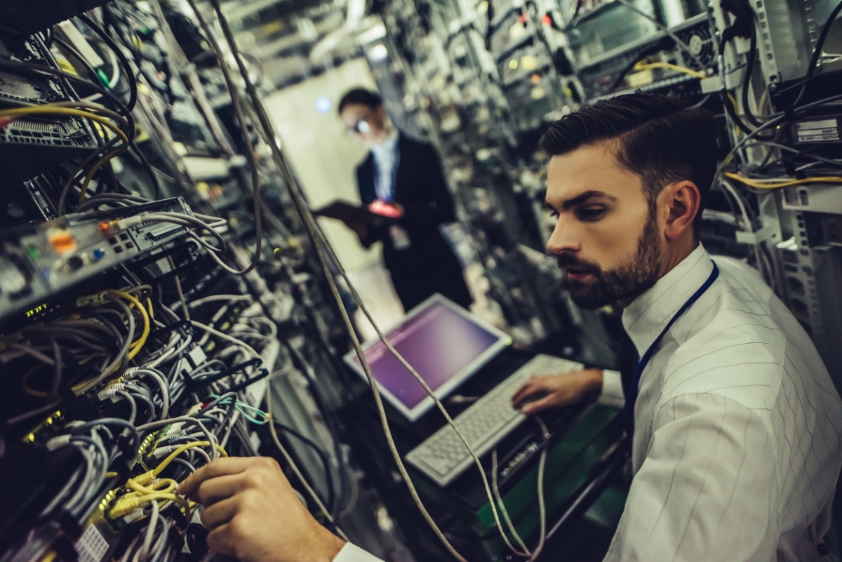 A man in a server room working on a computer and adjusting network cables, with another person working in the background