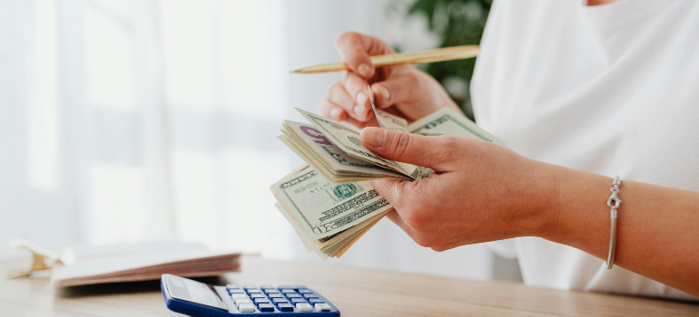 Person holding and counting dollar bills, with a calculator on the table.