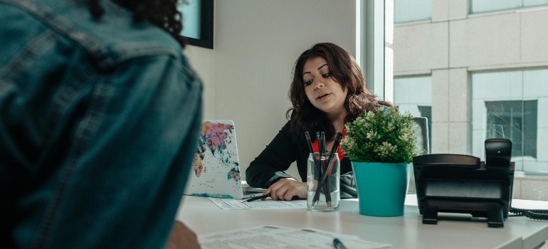 A businesswoman is holding a pen and sitting at her office, talking about movers in Reston to her client.